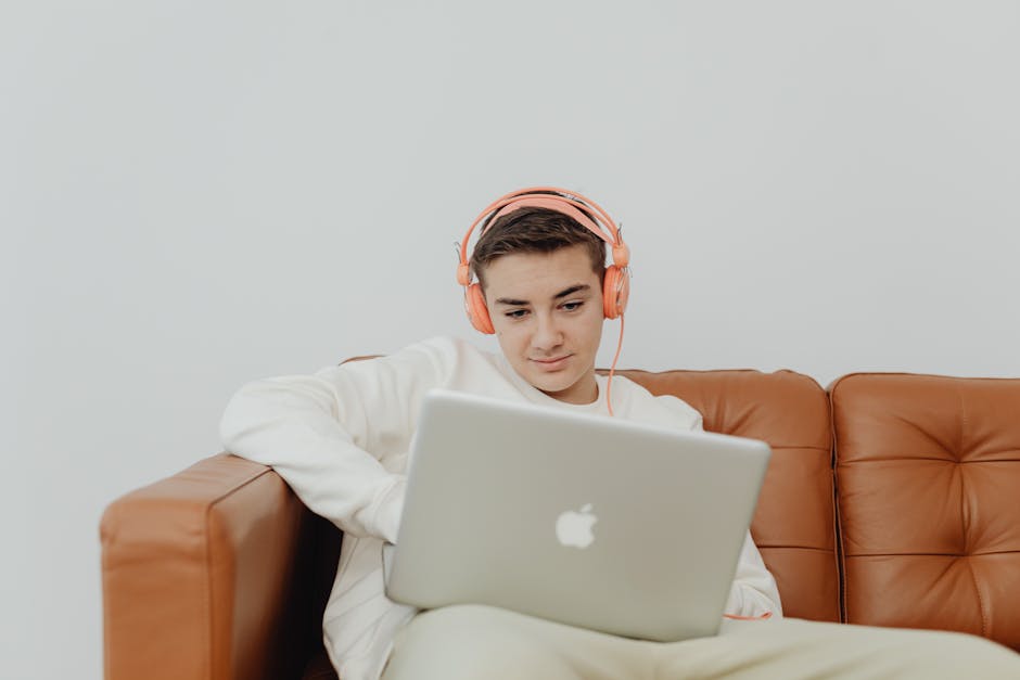 Young man sitting on a couch using a laptop with headphones, enjoying music indoors.